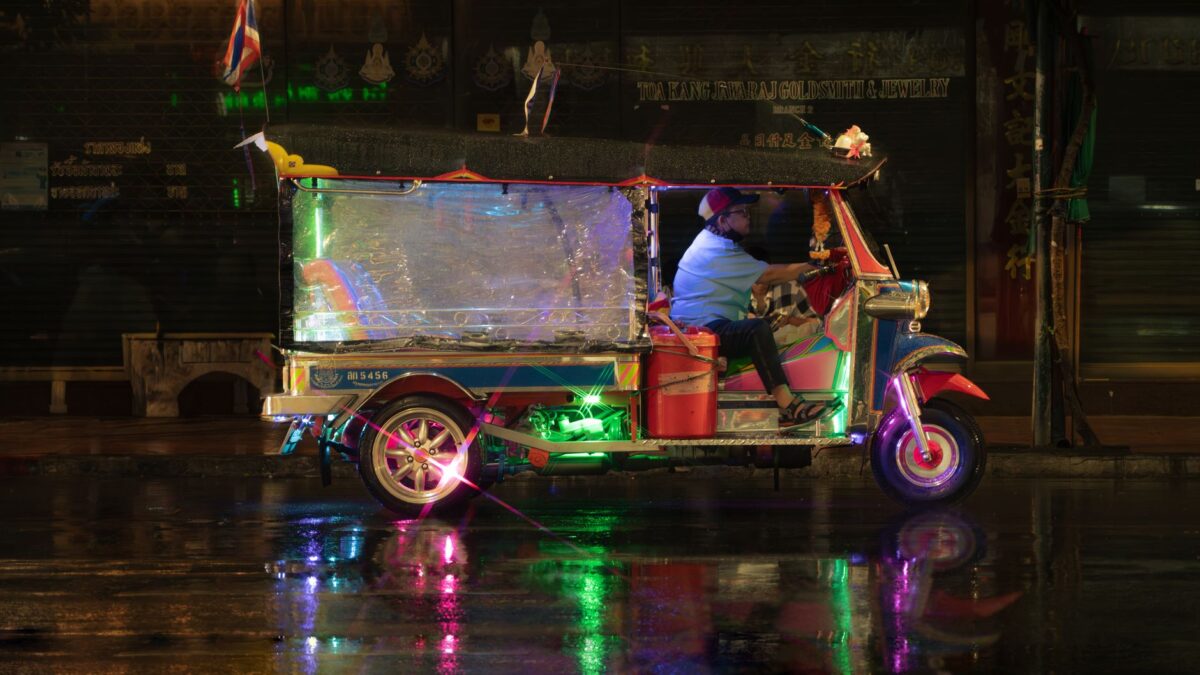 A vibrant tuk-tuk drives through a rainy Bangkok street, reflecting colorful lights.