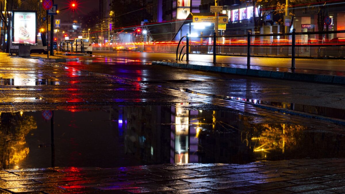 Urban night scene with vivid reflections on wet pavement, highlighting city lights and rainy atmosphere.