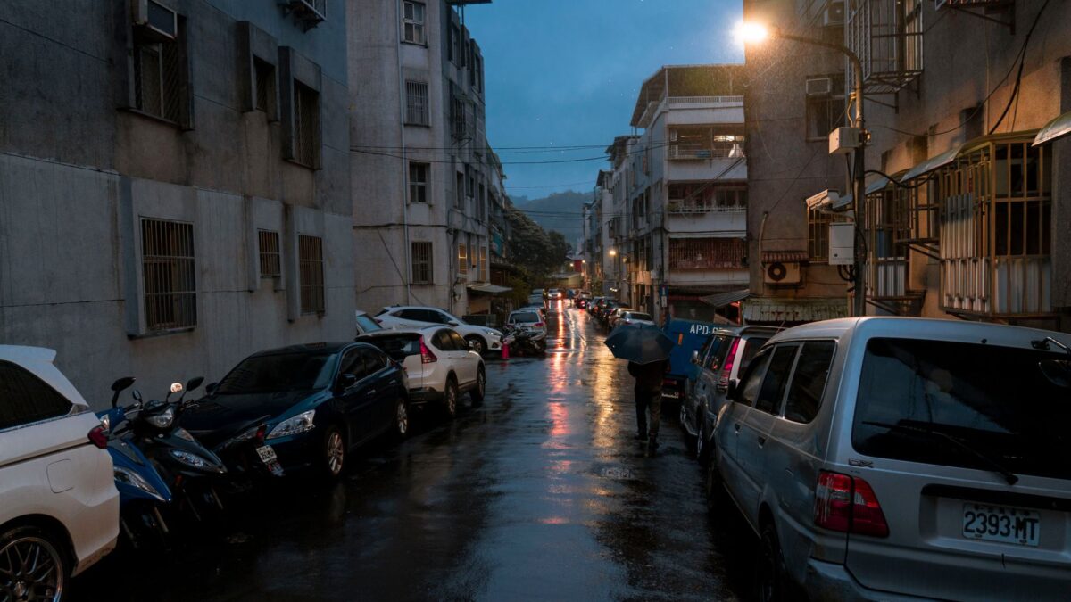 Moody urban street during a rainy night with reflections, parked cars, and streetlights.