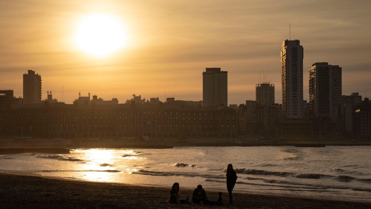 Silhouetted figures on Mar del Plata beach at sunset with city skyline in view.