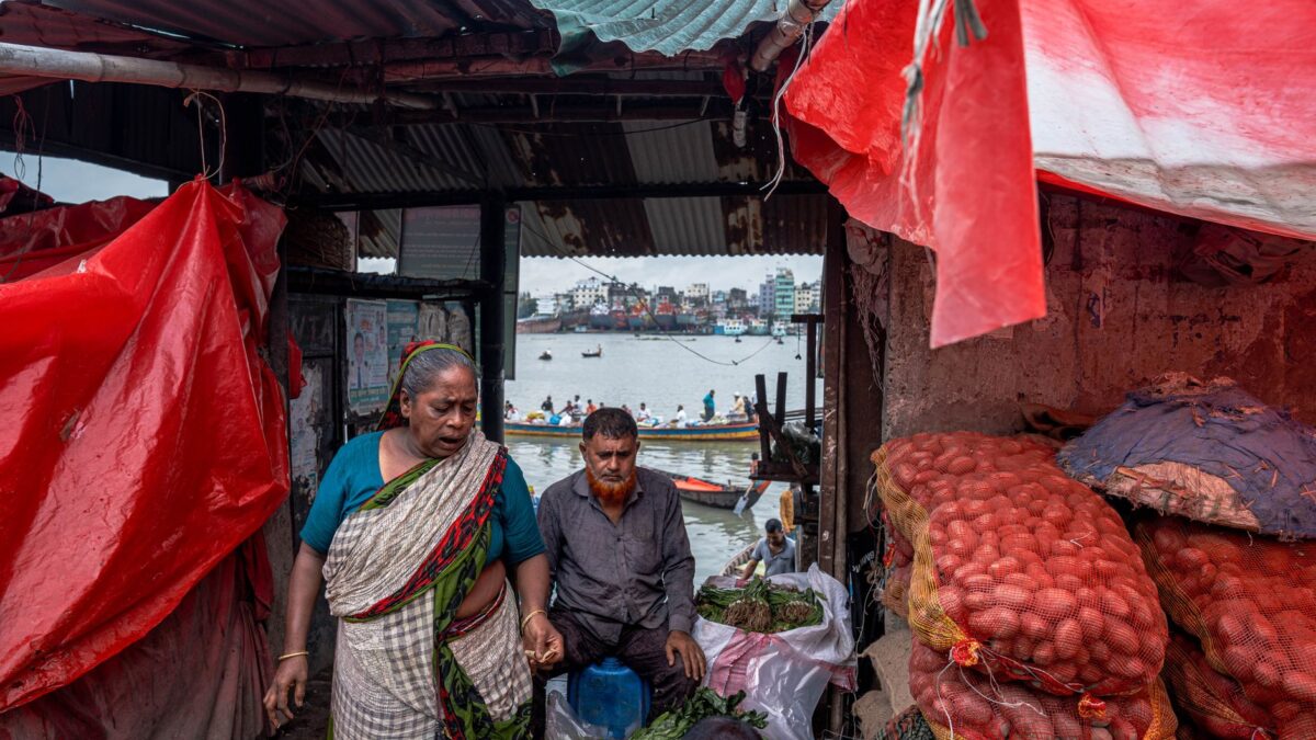 Vibrant outdoor market scene by a river with vendors selling fresh produce under colorful tarp.