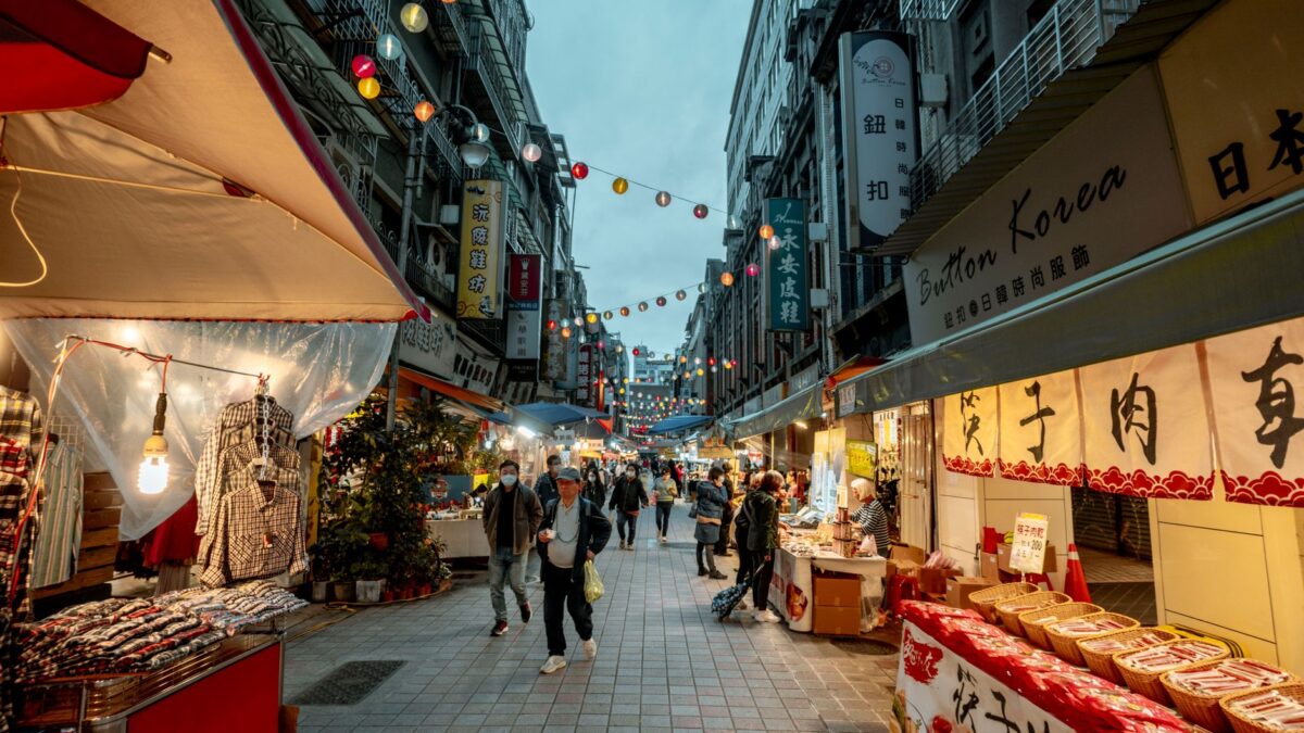 Busy street market in Asia at dusk, featuring shops, lanterns, and people shopping.