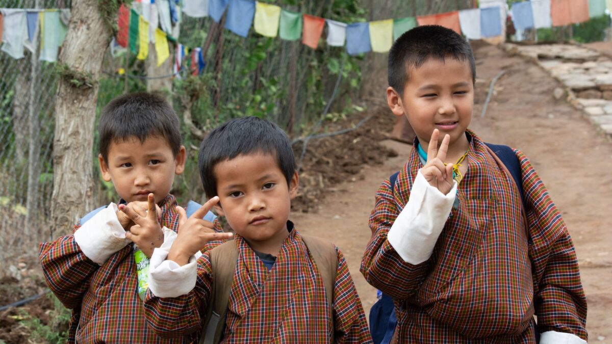 Three Bhutanese boys in traditional attire posing with peace gestures outdoors on a path with prayer flags.