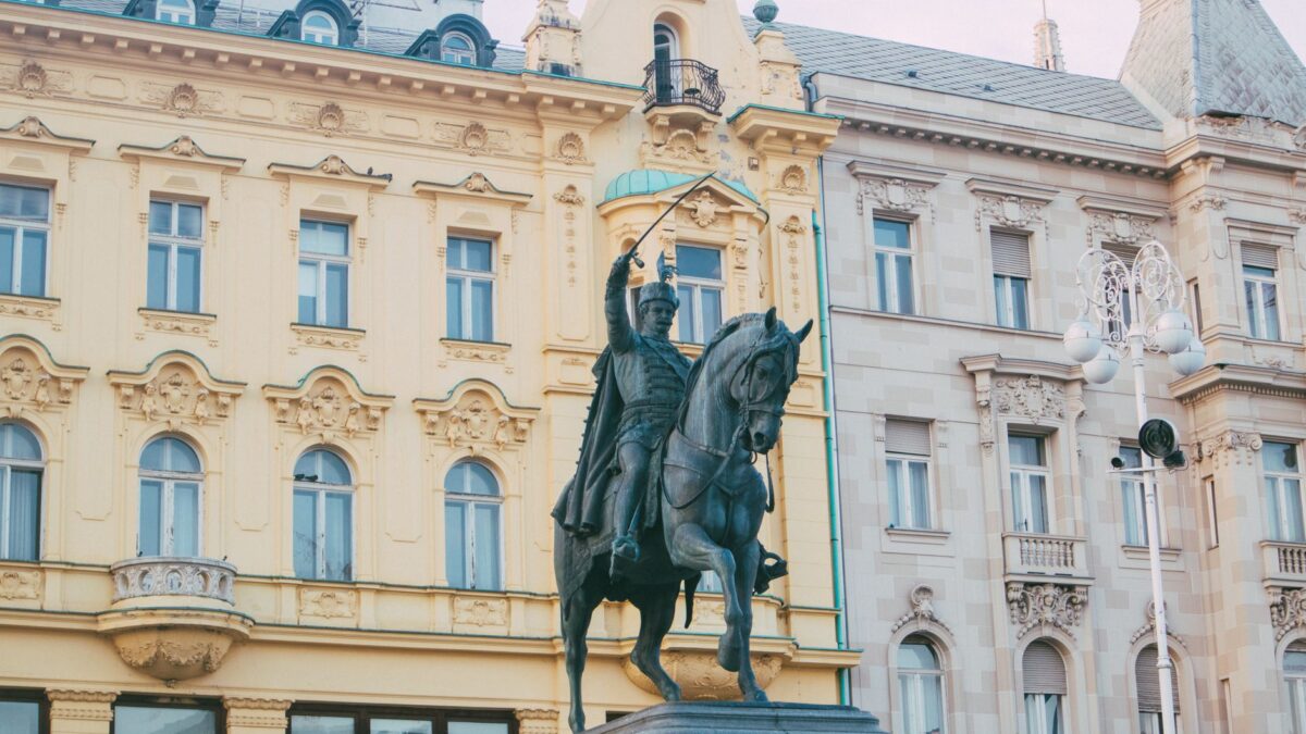 Historic equestrian statue in front of yellow and ornate townhouses in urban area.