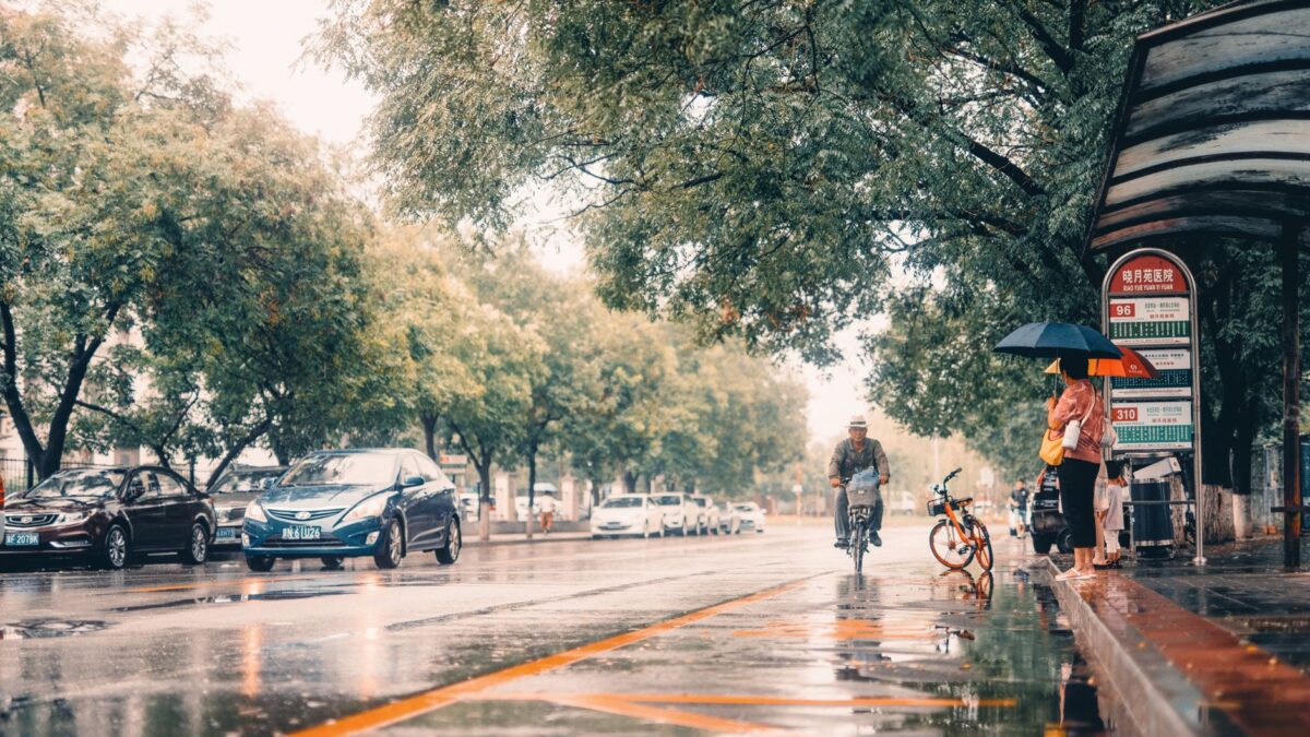 People wait under umbrellas at a Beijing bus stop while a cyclist rides by on a rainy day.