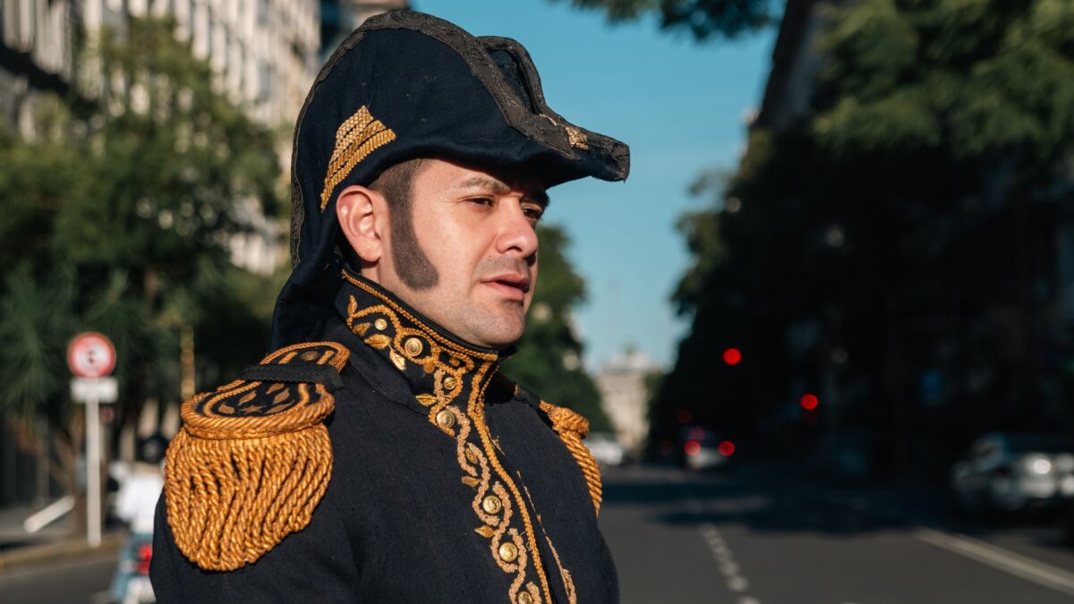 Adult male wearing elaborate historical military uniform standing proudly in a sunlit urban environment.