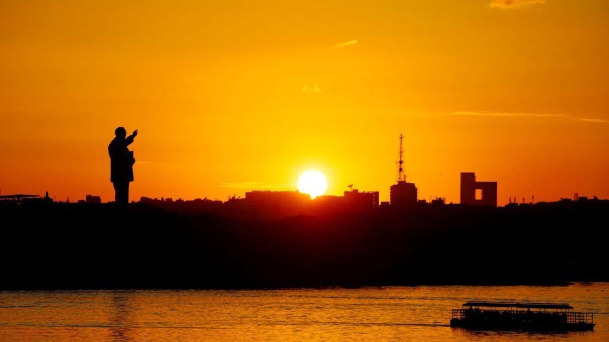 Silhouette of city skyline and boats on river at sunset, with vibrant orange sky.