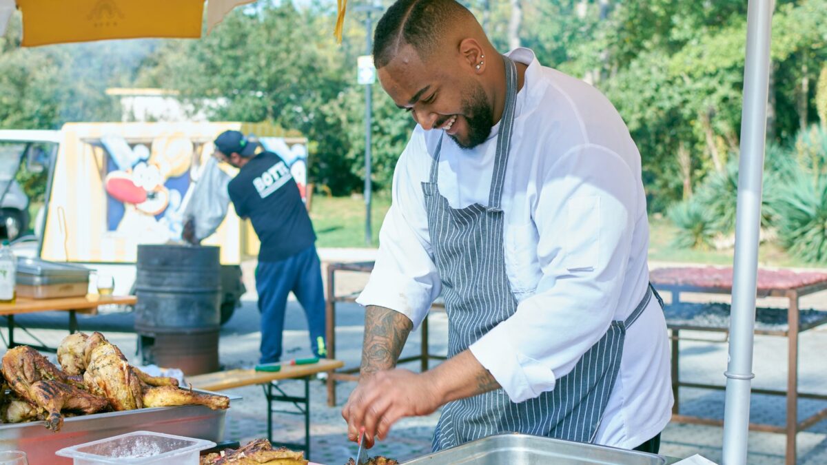 Happy chef preparing grilled chicken at an outdoor food market stand in a sunny setting.