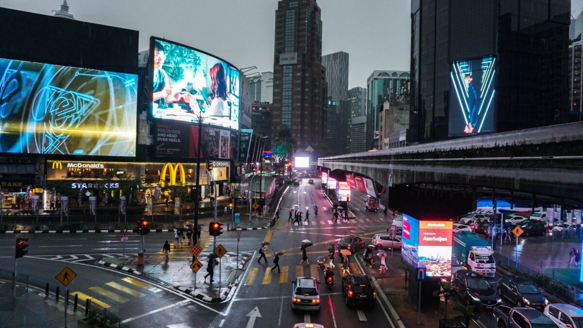 Crowded intersection in Kuala Lumpur at night, with vibrant billboards and traffic.