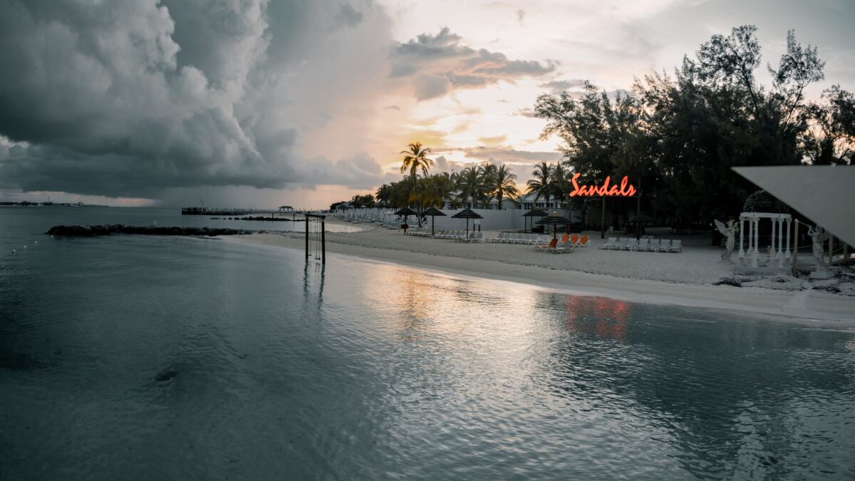 A serene beach resort at sunset with dramatic clouds and palm trees reflecting over tranquil waters.