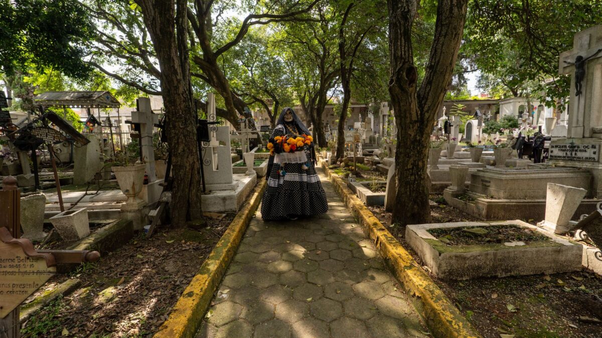Catrina figure in a cemetery holding flowers, representing Mexican Day of the Dead traditions.