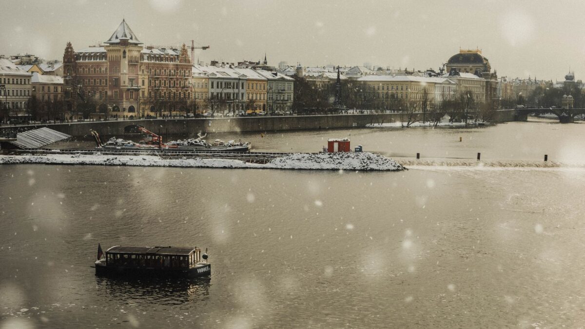 A tranquil winter scene of a snowy riverfront with a lone boat and historic buildings in the background.