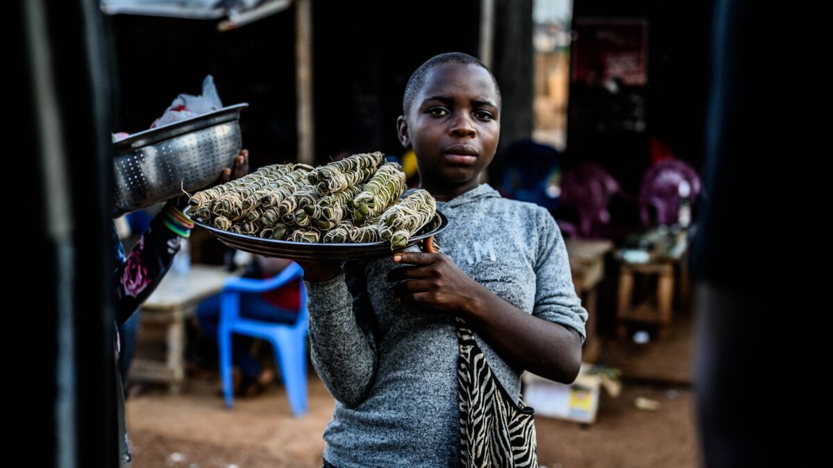 A young vendor presenting grilled sticks at an outdoor market setting.